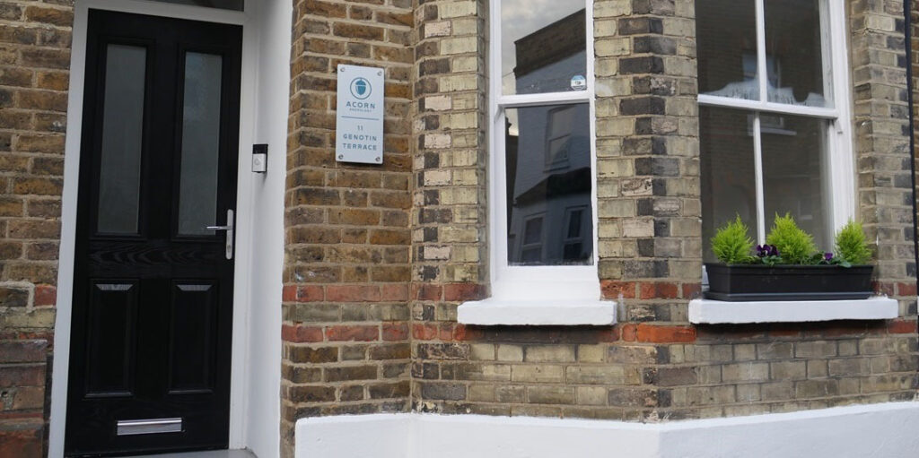 Entrance of a brick building with a black front door, white-framed windows, and a wall sign reading “Acorn, 11 Genotin Terrace.”